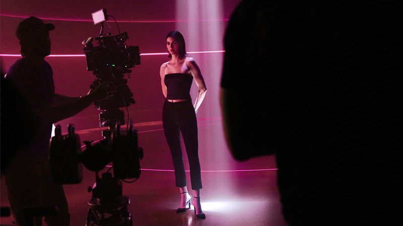 Modelo posa em estúdio rosa enquanto equipe de filmagem grava com câmera em primeiro plano.