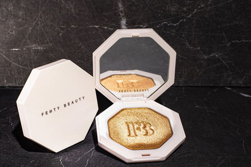 Luxury product photo of a Fenty Beauty highlighter compact open to reveal a shimmering gold powder against a dark marble background.