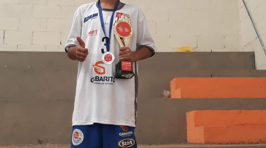 A imagem mostra um adolescente em pe, mostrando medalha que recebeu apos competicao