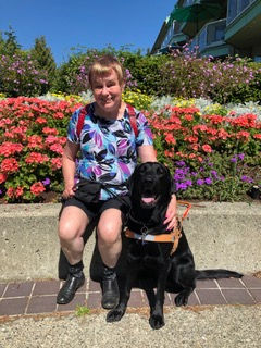 Betty with her guide dog Mac sitting in front of a bed of flowers