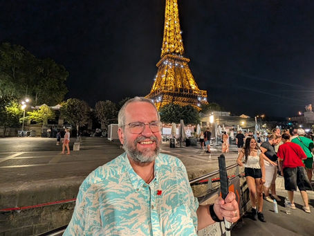 Richard, smiling and white cane in hand stands in front of the Eiffel Tower lit up at night