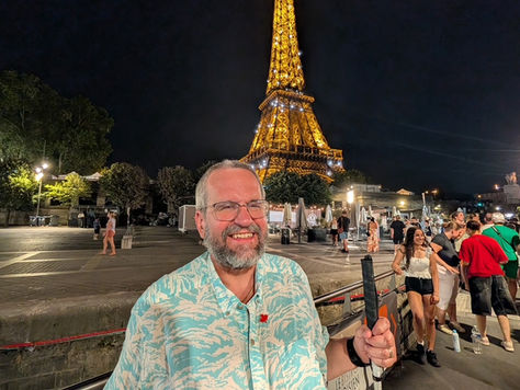Richard, smiling and white cane in hand stands in front of the Eiffel Tower lit up at night