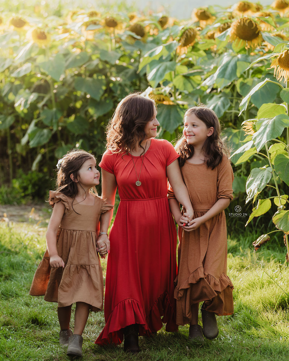 mother holds hands of each daughter on either side of her while walking in a sunflower field