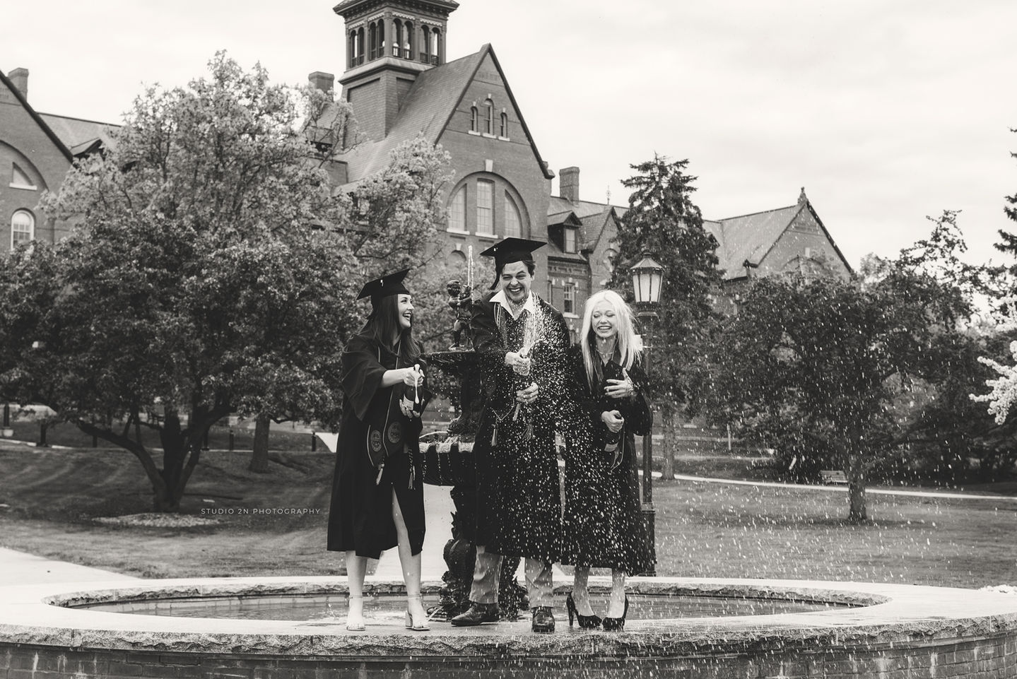 black and white photo of three college students standing on a campus fountain popping champagne