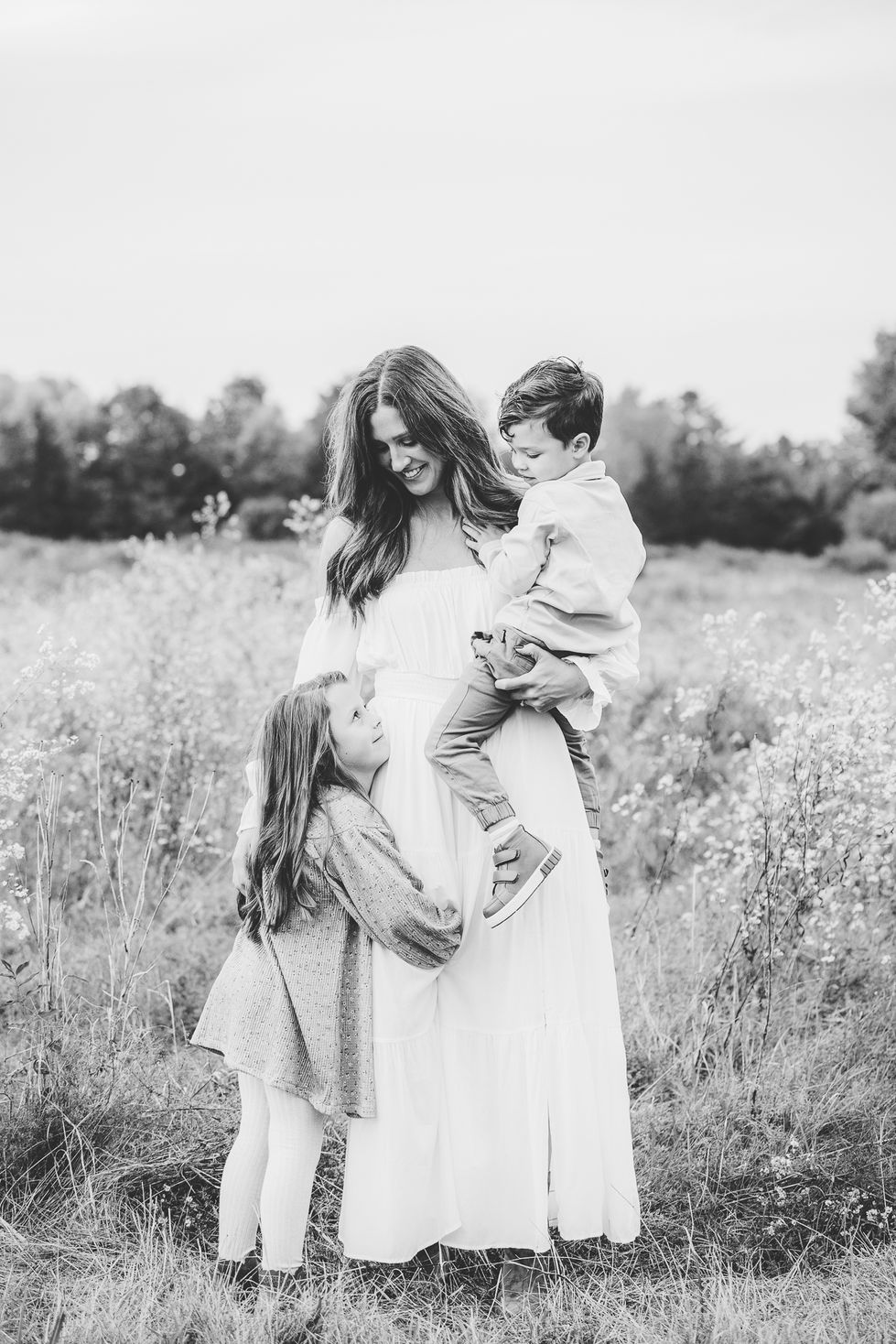 mother standing in grassy field holding young son and looking down at young daughter who is hugging her leg