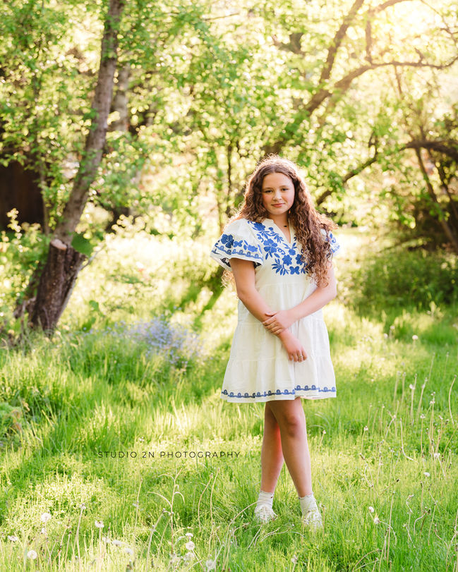 tween girl with brown curly hair in a white dress with blue detailing standing in a grassy area surrounded by trees