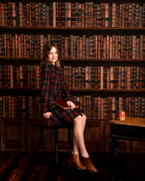 tween girl in a red plaid dress sitting on a stool holding a book in front of a library wall 