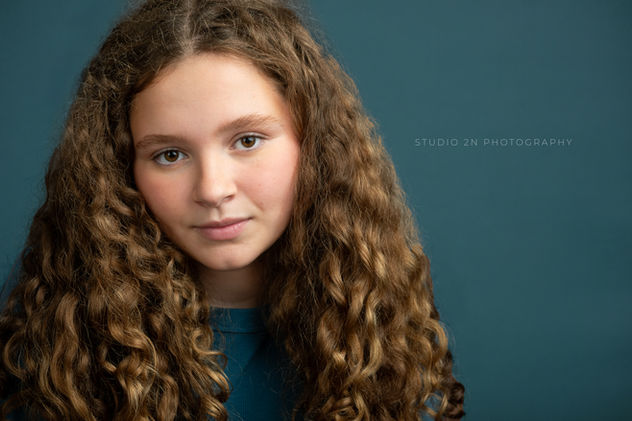 child actor headshot of tween girl with dark blonde, curly hair on a teal background