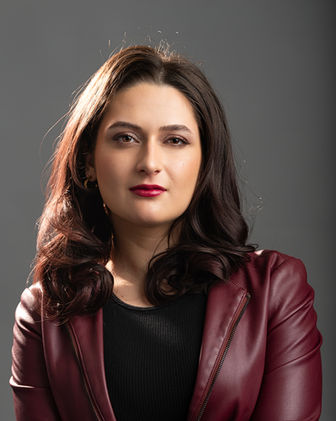 acting headshot of young middle eastern woman with brunette hair wearing a maroon leather jacket on a gray background