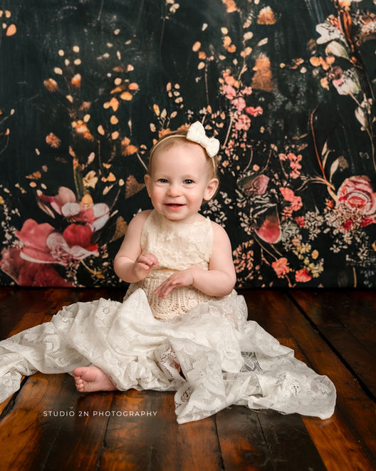 infant girl in cream dress and white bow sitting on a wooden floor with a dark floral background