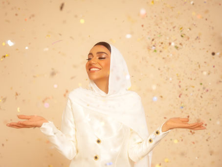 امرأة ترتدي حجابًا أبيض تبتسم برضا، محاطة بقصاصات لامعة ومتناثرة في الخلفية. الأجواء مرحة ولطيفة.