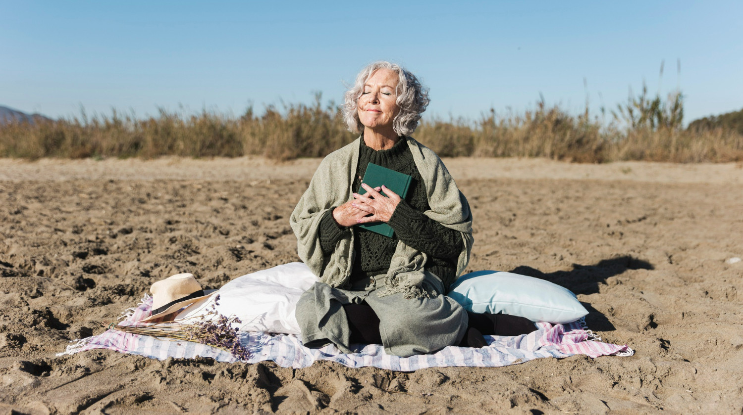 Gray-haired woman meditates on a sandy beach, holding a green book, seated on a blanket with pillows under a clear blue sky.