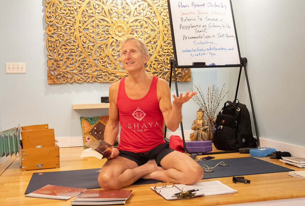 Smiling yoga instructor in red tank top sits cross-legged on a mat, gesturing beside a whiteboard with handwritten notes.