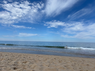 calm waves at the beach, blue sky with wispy white clouds, tan colored sand, a person swimming in the waves