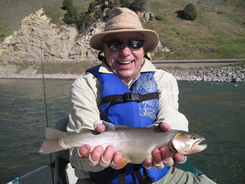 A smiling man in a wide-brimmed hat and a blue life jacket holds a trout with both hands. Rocky land rises behind him.