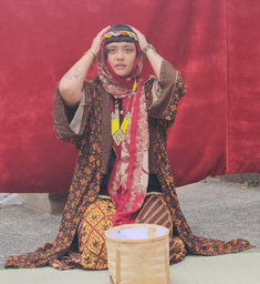 Person in patterned attire sits on the ground, hands on head, with a red background. Wicker basket nearby adds to traditional feel.