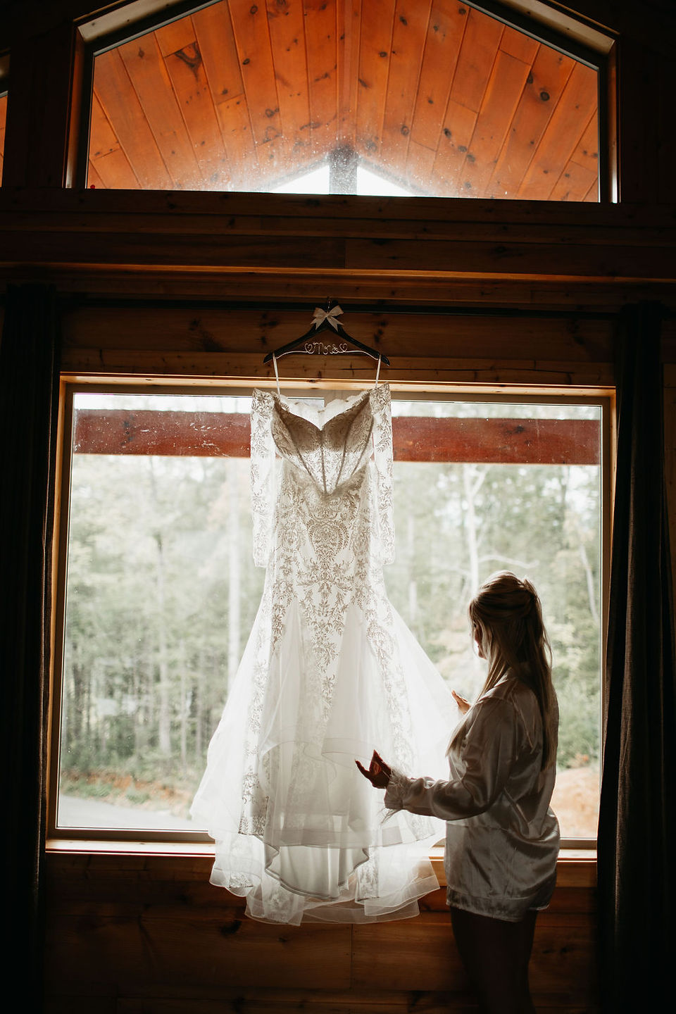 bride standing in the window of her bridal suite with her wedding dress hanging in the window