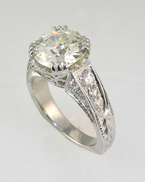 Vintage-style engagement ring with a round center diamond in a three-claw prong setting, filigree gallery, milgrain edges, engraving, and graduated side diamonds.