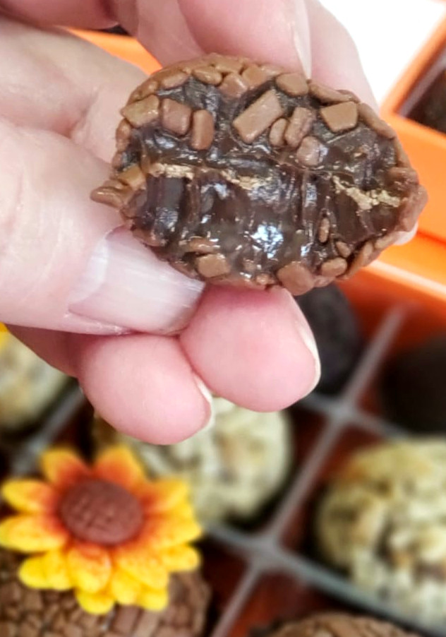 Brigadeiro artesanal mordido sendo segurado, com caixa de brigadeiros ao fundo.