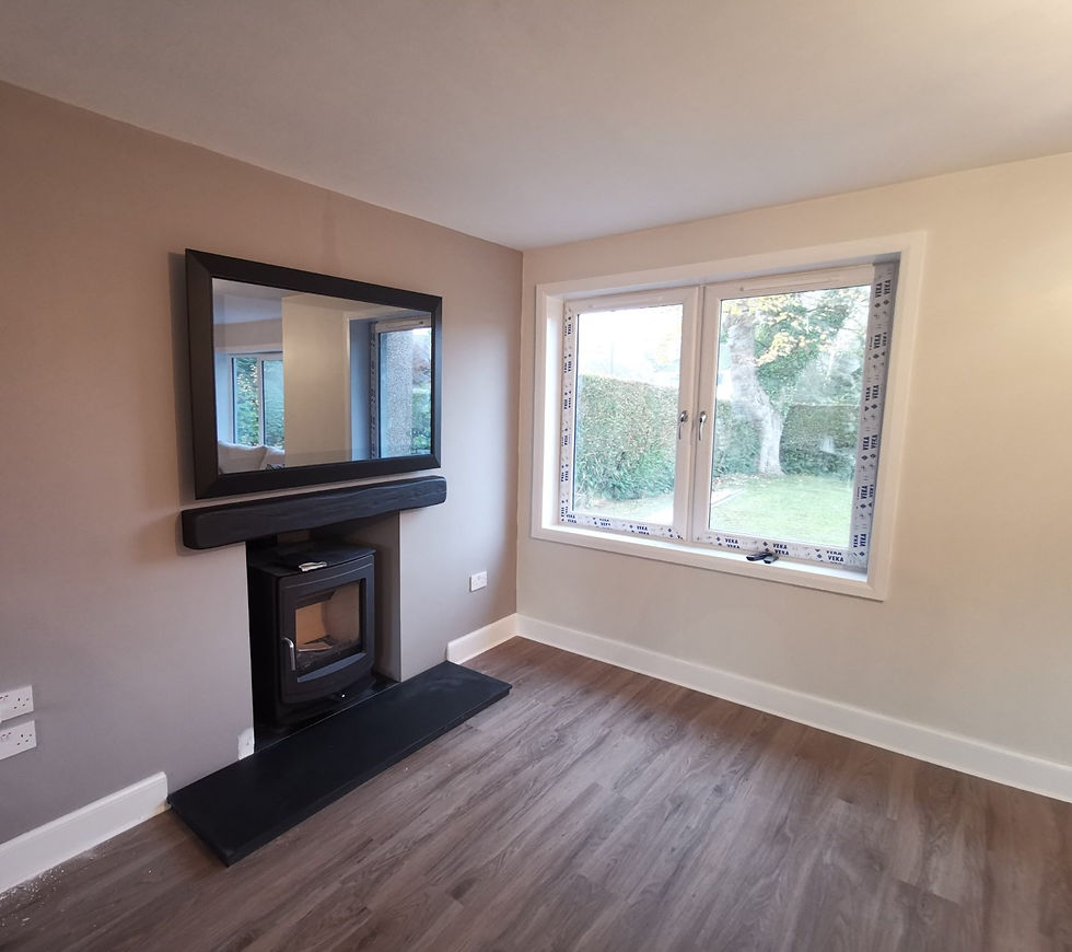 A black framed mirror tv is installed above a black fireplace with a black wood burner in it. The walls are white and there is a large window at the right hand side. The flooring is wooden and the house is fairly bare as just renovated.