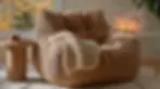Cozy tan corduroy bean bag with beige blanket. Wooden table with a terracotta cup beside it. Autumn scene outside window, candles lit inside.