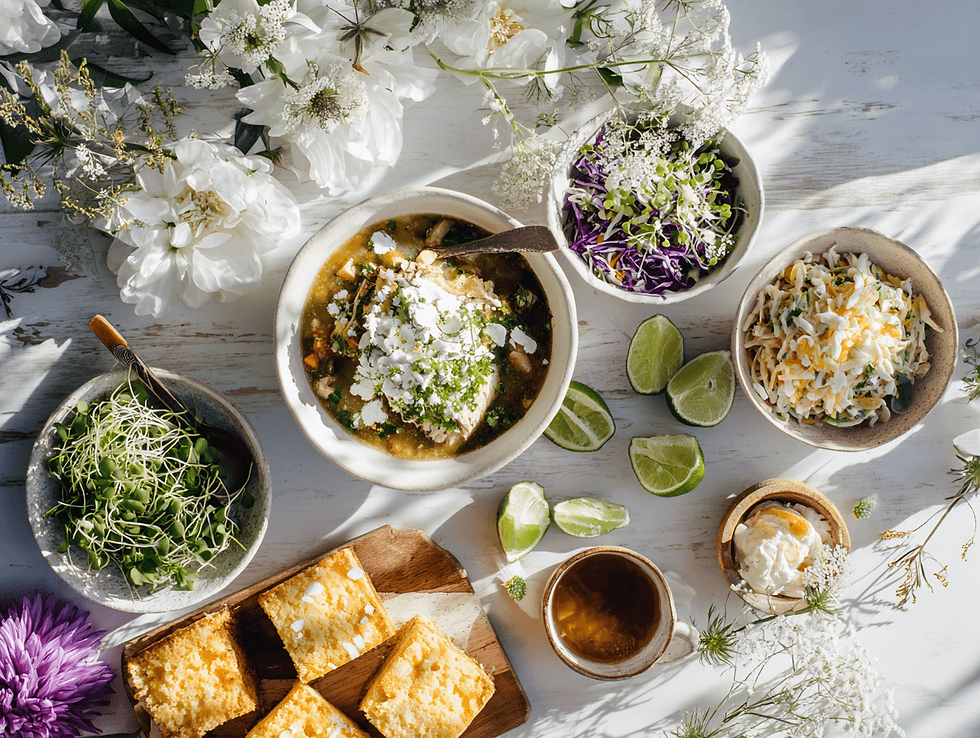 Top-down scene of a bright chili spread with a bowl of green chili, fresh herbs, shredded cabbage, limes, microgreens, and sliced cornbread arranged with white flowers on a sunlit table.
