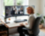 Smiling woman in a home office on a video call across three monitors, seated at a desk by a bright window.