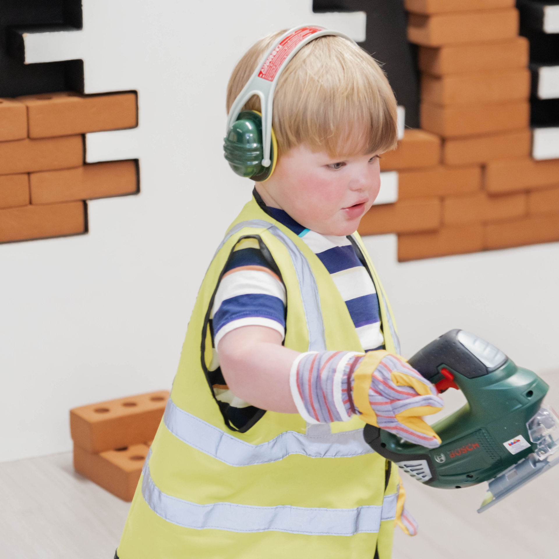 young boys dressed in a hi vis and ear defenders carrying a power tool at petite street play village