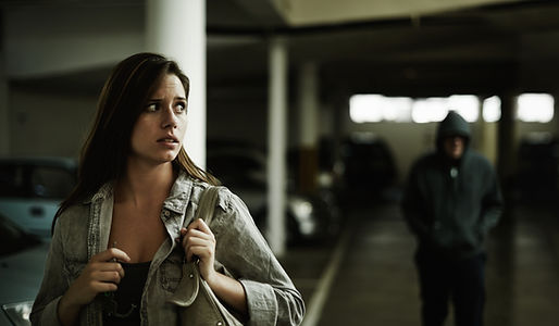 Uma mulher andando em uma garagem de um edificio. Ela está com um olhar assustado, porque atrás dele vem um homem com uma jaqueta, de capuz, e está com o capuz na cabeça e as duas mãos mno rosto. Ele está andando na direção da mulher. Atrás aparece muitos carros estacionados.