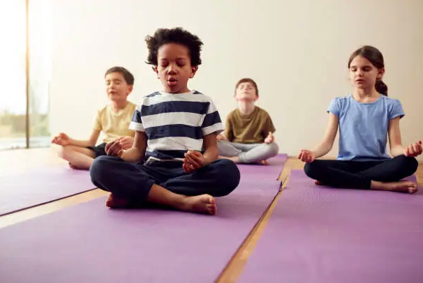 Four children sit cross-legged on purple yoga mats, meditating in a bright studio, calm and focused.