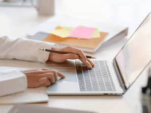 Person in white shirt typing on a laptop at a desk with a notebook and colorful sticky notes. Bright, calm workspace with plants.