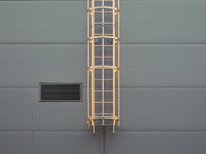 Metal ladder with a protective cage on a gray corrugated wall. Vent with horizontal slats on the left.
