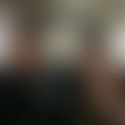 Smiling man and woman pose with thumbs up in a store. Man wears a cap, apron, and name tag. Woman wears green beads. Bright, busy background.