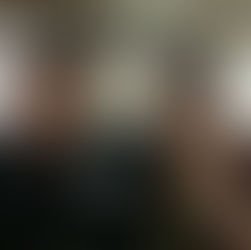 Smiling man and woman pose with thumbs up in a store. Man wears a cap, apron, and name tag. Woman wears green beads. Bright, busy background.