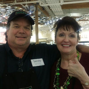 Smiling man and woman pose with thumbs up in a store. Man wears a cap, apron, and name tag. Woman wears green beads. Bright, busy background.