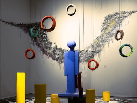 Kunstinstallation mit blauer Figuren-Skulpzur, hängenden bunten Ringen und gelben Zylindern im Ausstellungsraum