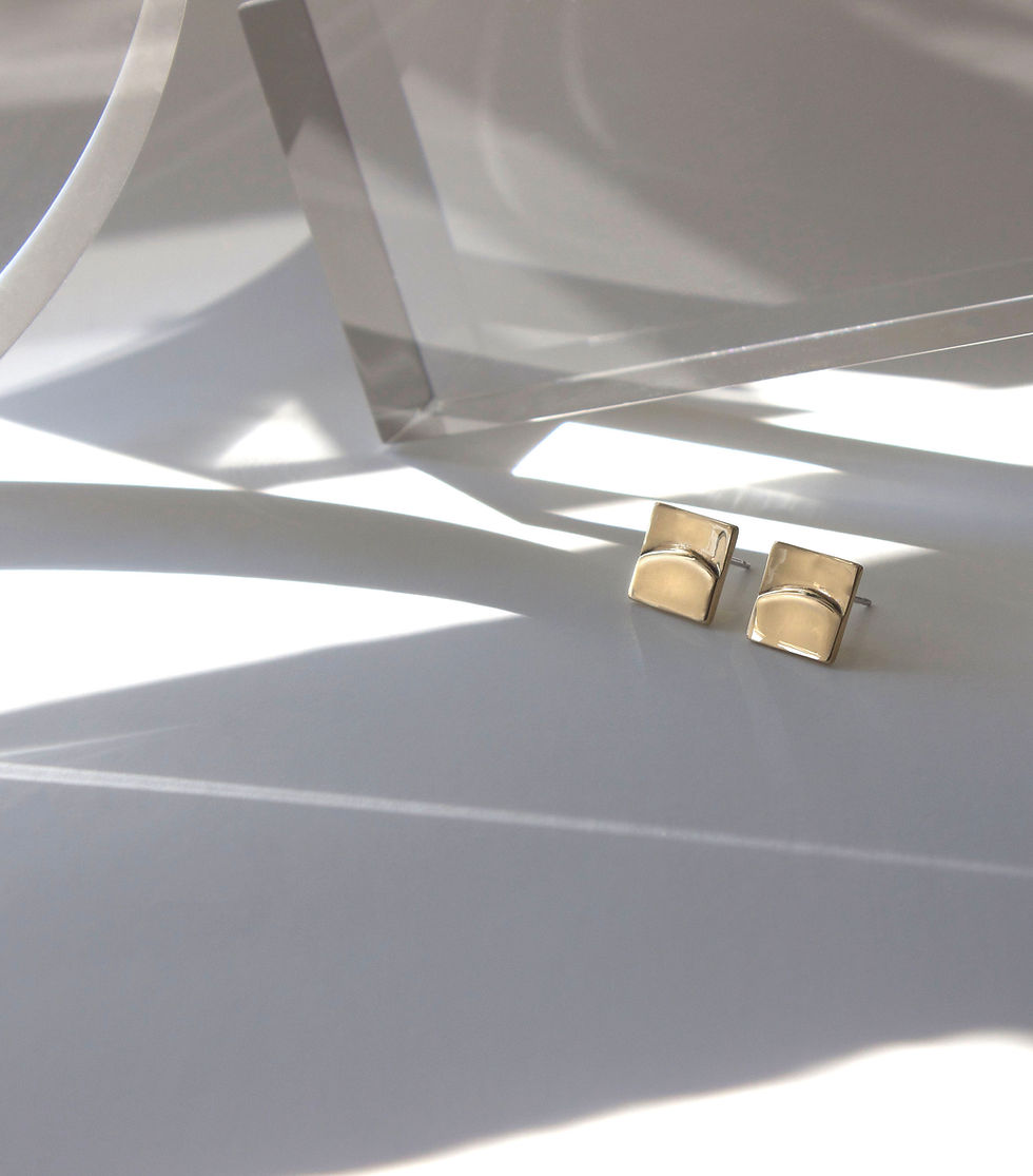 Thumbnail: Gold tone square stud earrings featuring a raised half circle design.  Background is off white with streaks of light. Various clear resin props in the background. 