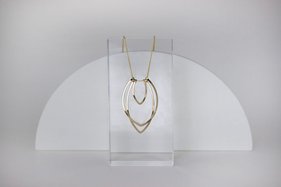 Thumbnail: Further perspective of Gold Fill Small Flame Necklace. A design of three tiers of upside down tear drop shapes varying from long to wide to small and center. Design is an abstract version of a flame. This is the small version of the design. 
Prop is a clear resin rectangle lighting is  bright and white. A white half circle is featured behind the clear resin prop. 