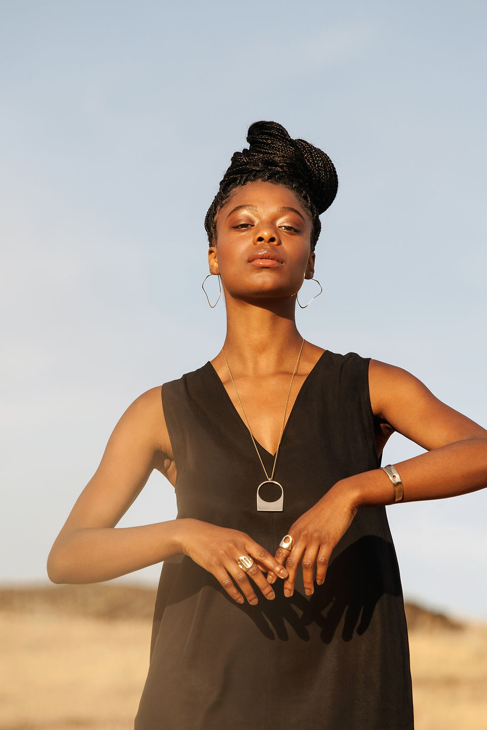 Thumbnail: Black Woman with long, black braided hair worn high up in a bun facing the camera, angling her arms almost in a bowing posture, the background feature soft yellow hills and blue sky. She is wearing a short sleeved, black, loose top and various gold tone jewelry.