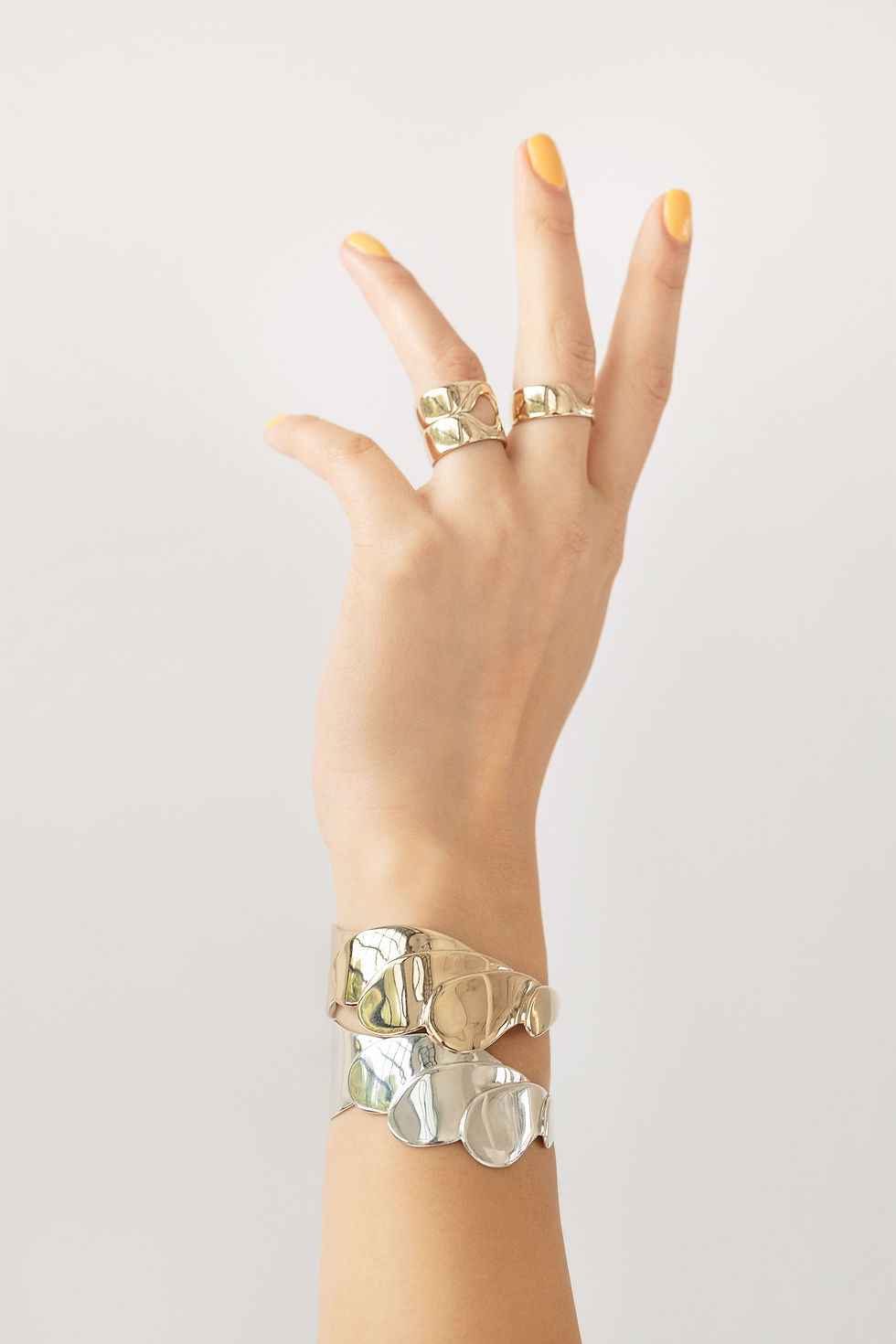 Thumbnail: Left hand raised with silver and gold tone cuffs on wearing multiple rings in silver and gold tone. 