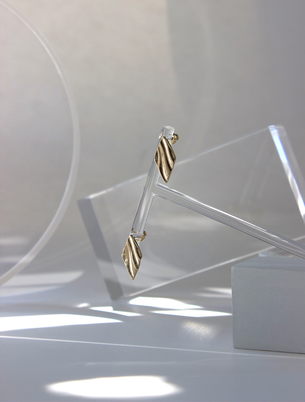 Thumbnail: Gold tone diamond shape earrings with deep bark light groove designs in the face of the metal, hanging from a clear resin earring stand with various clear resin props in the background. 