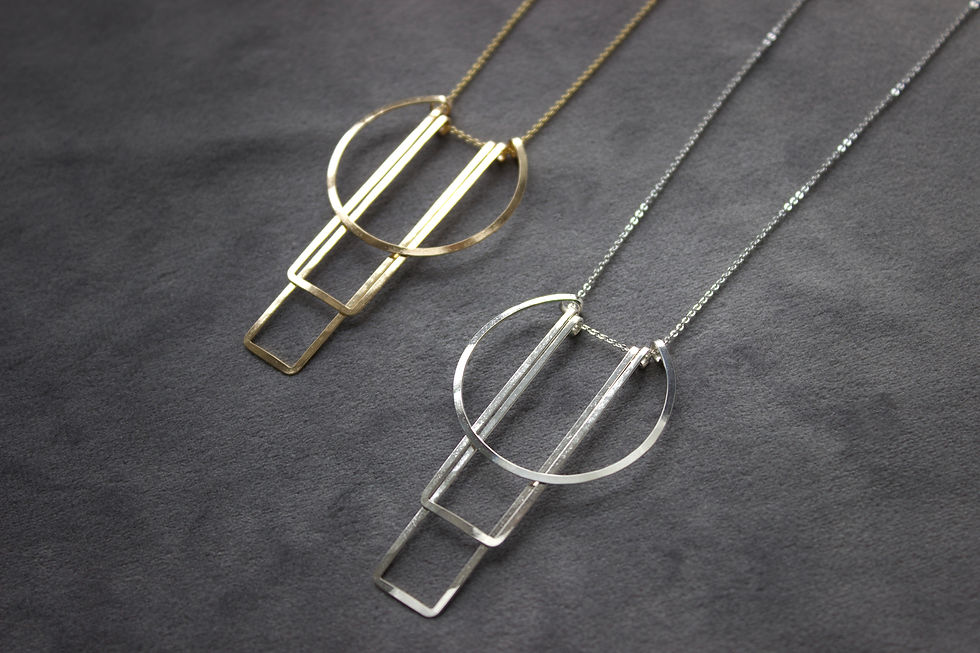 Thumbnail: Silver and gold geometric necklaces on gray velvet background with muted light. The muted light makes the metal and chain glimmer.