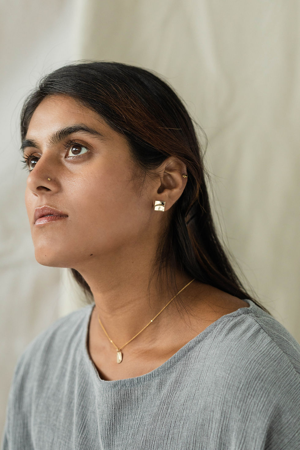 Thumbnail: Profile view of South Asian woman with long dark hair, profile view.  Background is a soft fabric canvas. She is wearing a jean blue top. 