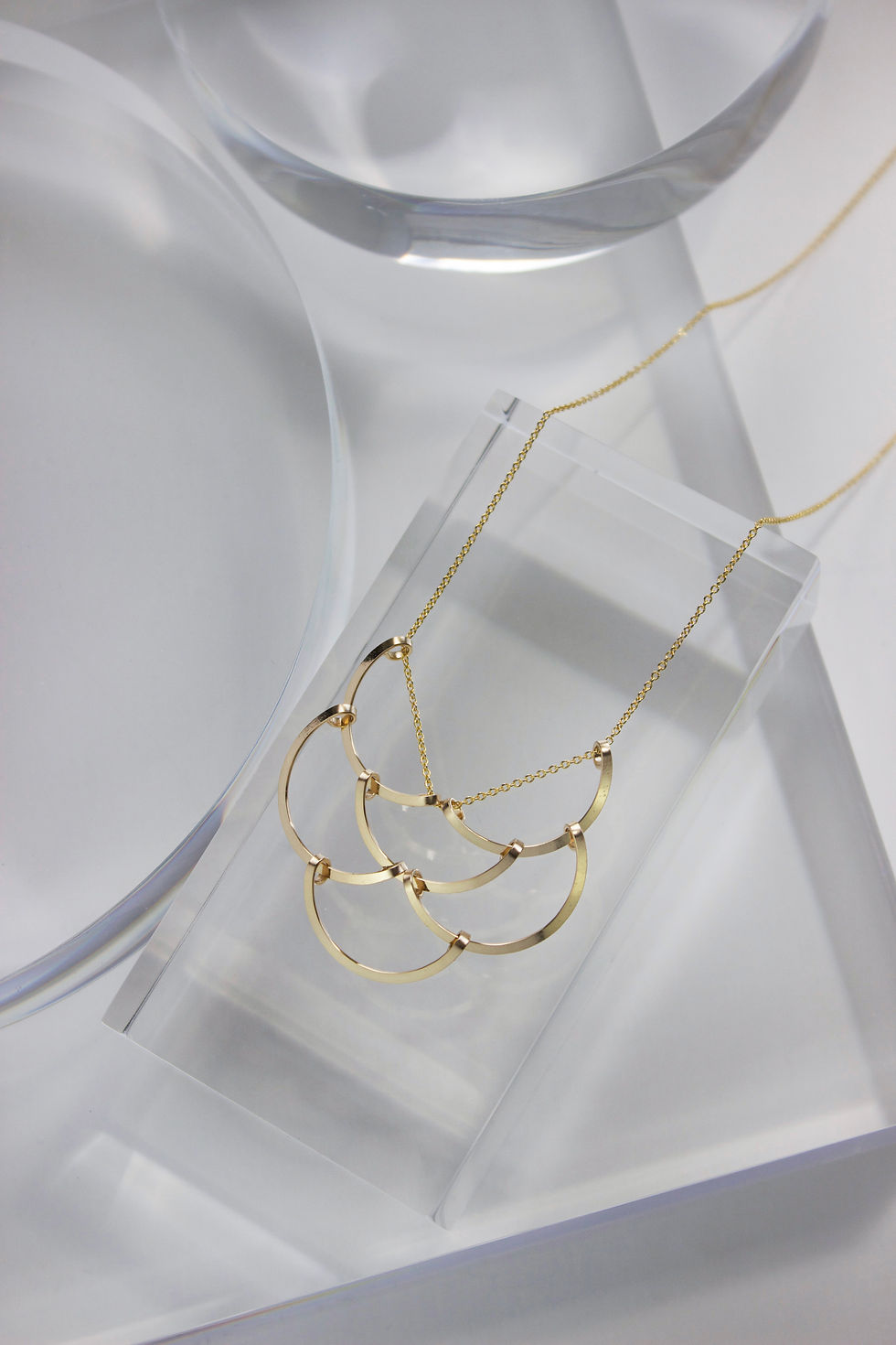 Gold fill necklace on various clear, resin geometric props. The necklace design features six Interwoven hammered gold wire crescents, creating a scale like effect.
