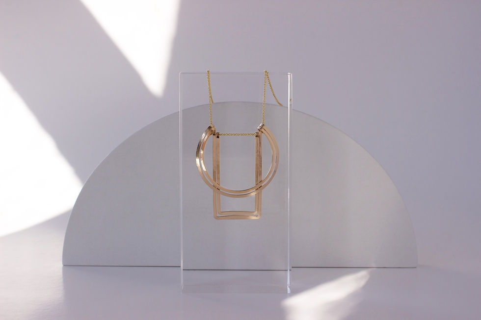 Thumbnail: Gold necklace on a clear, resin rectangle prop with a white wooden semi circle in the background the surrounding background color is bright lit with off-white shadows.