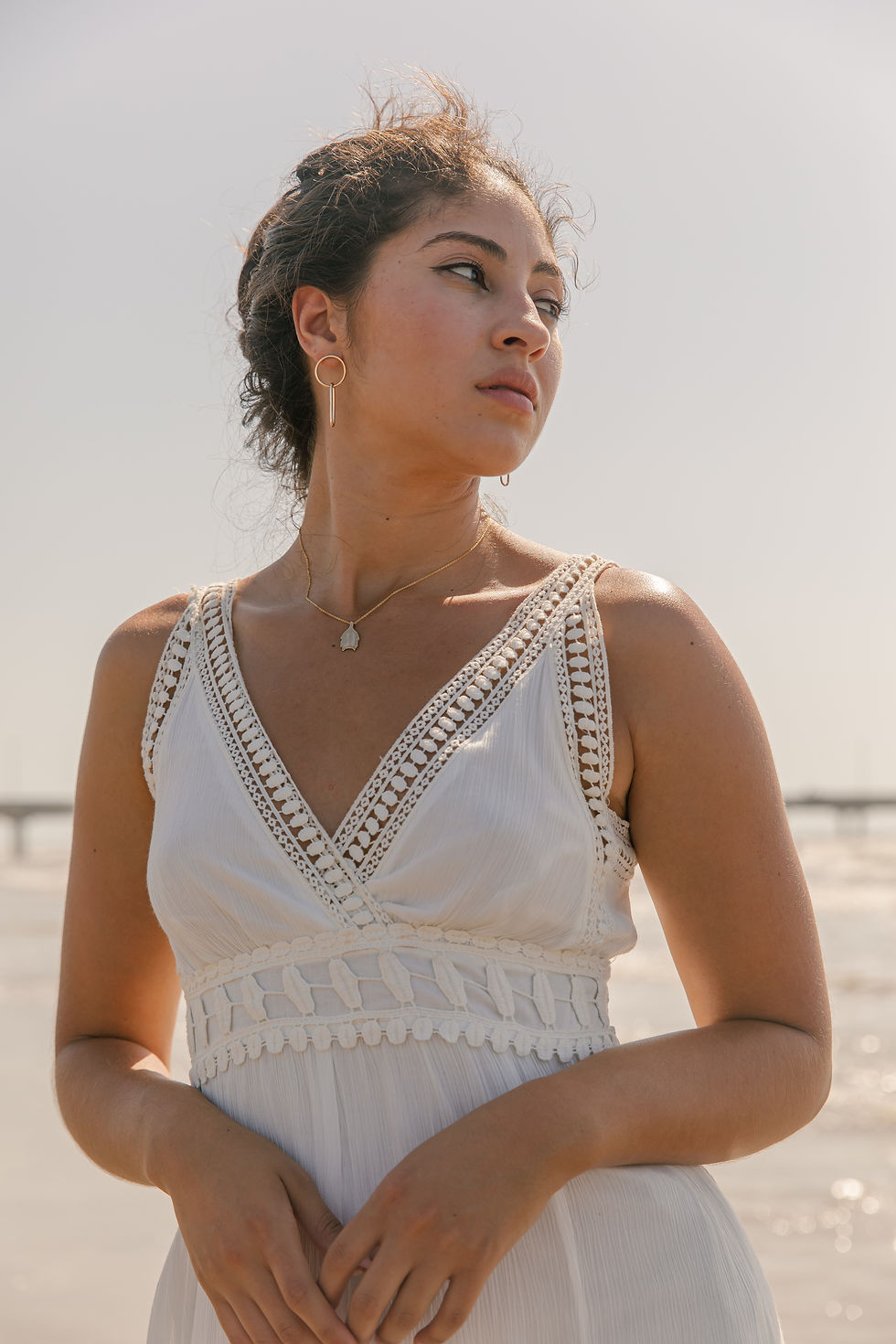 Thumbnail: Latina woman at the beach, looking over her left shoulder, ocean is in the background. She is wearing a white sleeveless dress and her brown hair is up in a bun. 