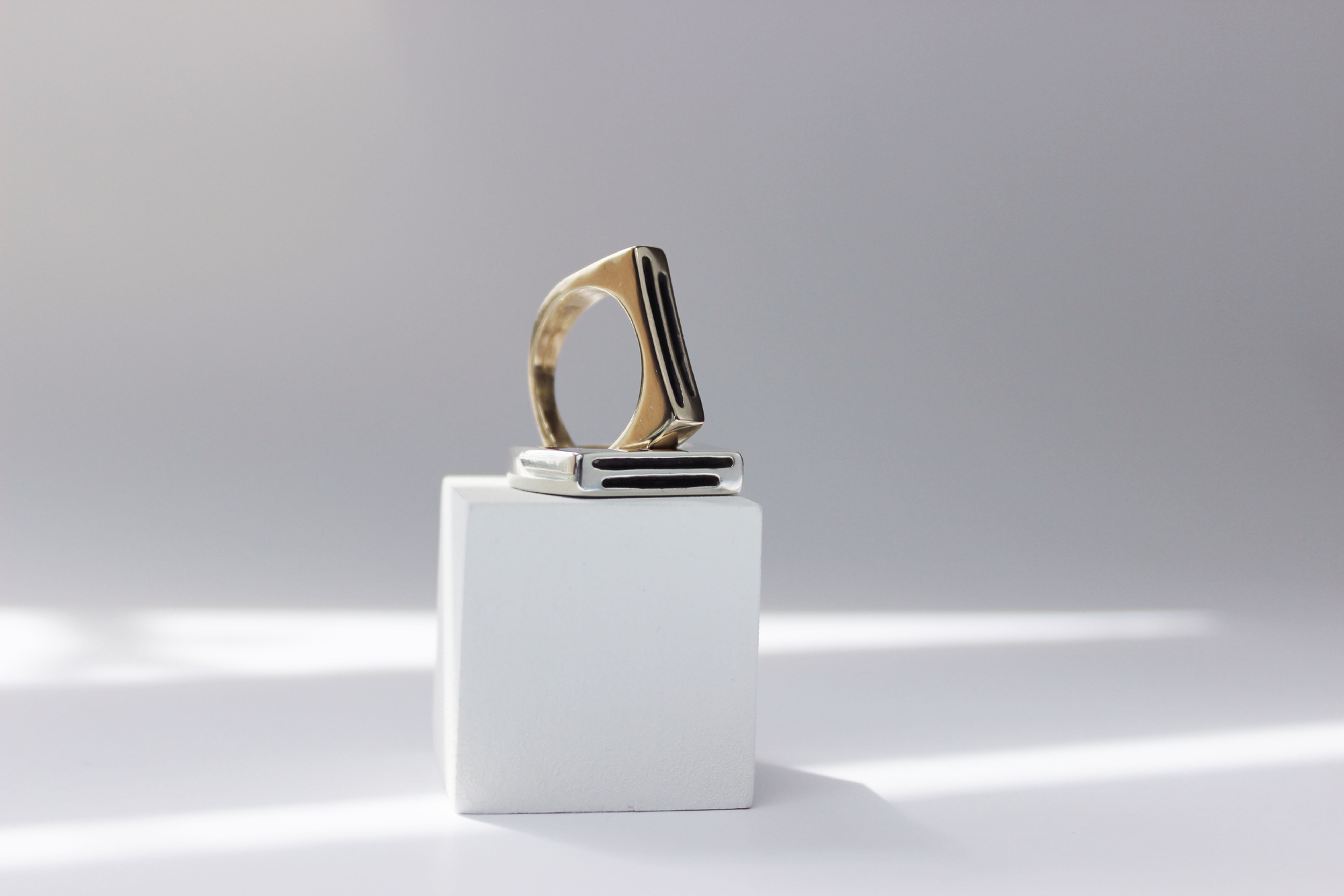 Silver and gold tone rings stacked on a white wooden square prop with dappled light in the background. Unison ring design features a signet style ring with two hollow lines running horizontally through the center of the flat top of the ring, one line slightly longer than the other but centered with the shorter line.