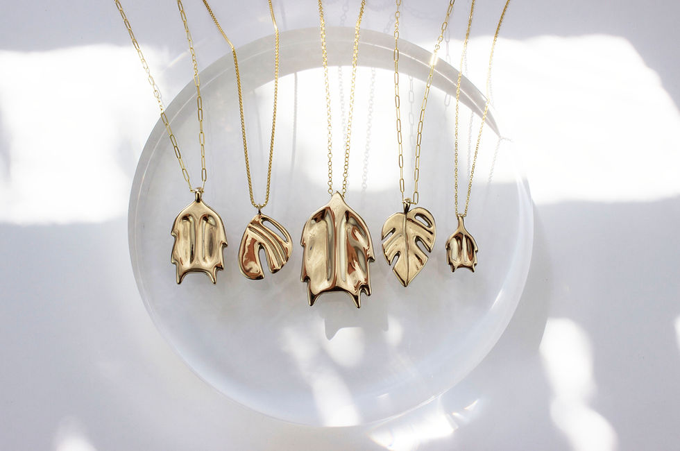 Thumbnail: A collection of gold plant inspired pendant necklaces on a clear round resin prop on a dappled light off white background.