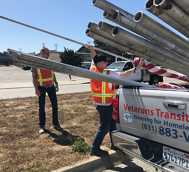 Two men in dark jeans and orange construction jackets grabbing flag poles out of the back of a silver pickup truck. The truck has text on the back. The red Text says Veterans Transitin center. The blue text below says housing for homeless veterans. Below that line reads (831) 883-VETS