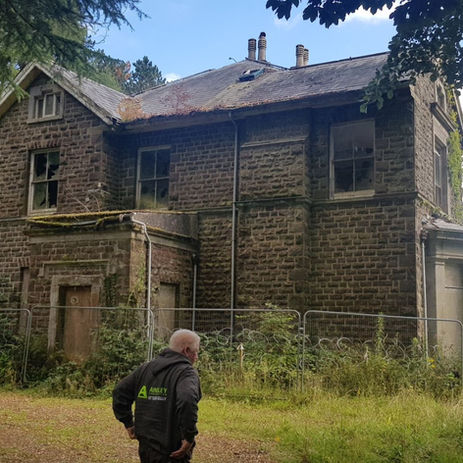 Surveyor in Ainsley jacket assessing a neglected stone manor behind security fencing.